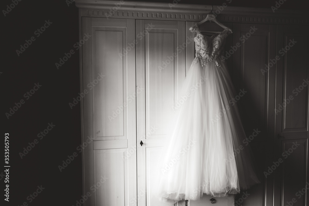 Black and white photo of wedding gown on hanger hanging on white wardrobe and waiting for bride. Wedding day concept.
