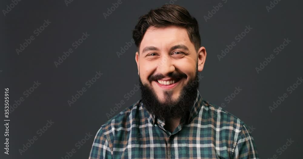 Close up portrait of handsome happy man with beard and mustache in checkered shirt smiling and winking to camera while standing on grey background indoor. Joyful funny Caucasian guy in good mood. Close up portrait of handsome happy man with beard and mustache in checkered shirt smiling and winking to camera while standing on grey background indoor. Joyful funny Caucasian guy in good mood.