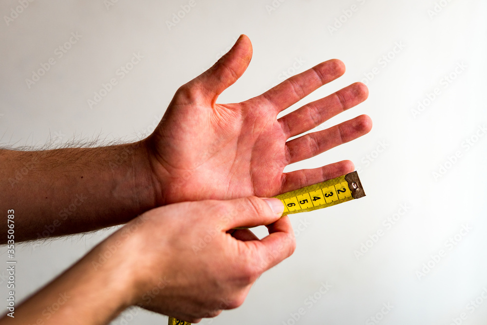 Person measuring the pinkie, little finger of his left hand with a yellow tape measure. White background. Olive skin tone.