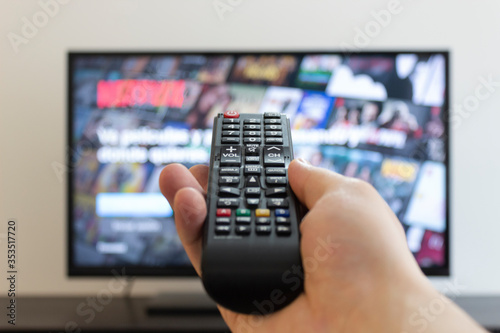 Closeup to a male hand holding a black TV controler with a blurred flat tv at background. indoor and entretainment concept photography 