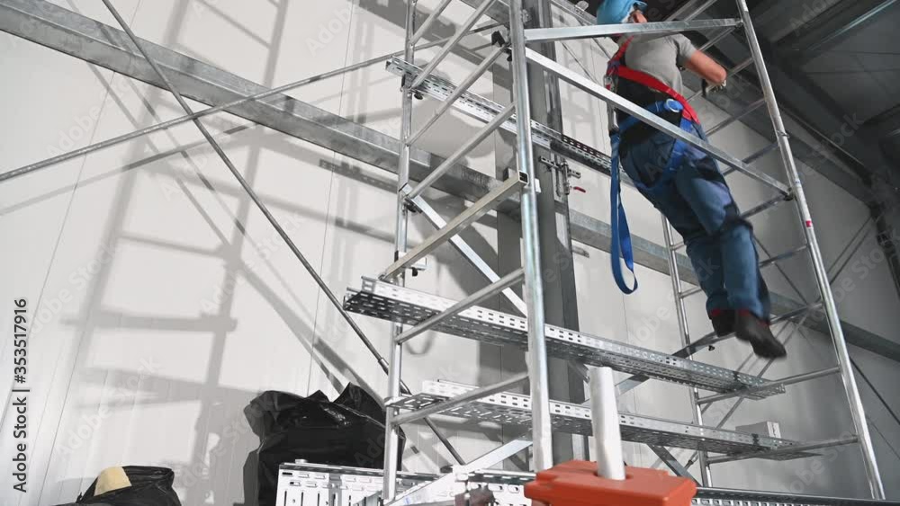 Caucasian Contractor Worker in His 30s Wearing Safety Harness Going Up Using Scaffolding To Replace Air Vent Elements Inside Warehouse Building.