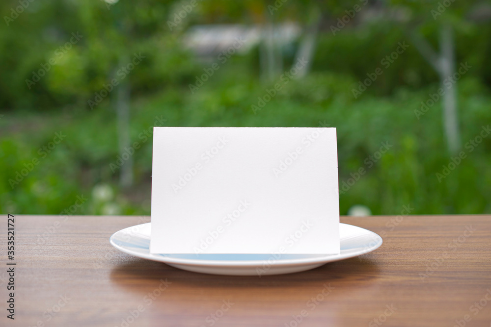 Blank standing bifold card on a classic empty white plate on the outdoor wooden table and green garden behind, clean template for you design presentation. Blank standing bifold card on a classic empty white plate on the outdoor wooden table and green garden behind, clean template for you design presentation.