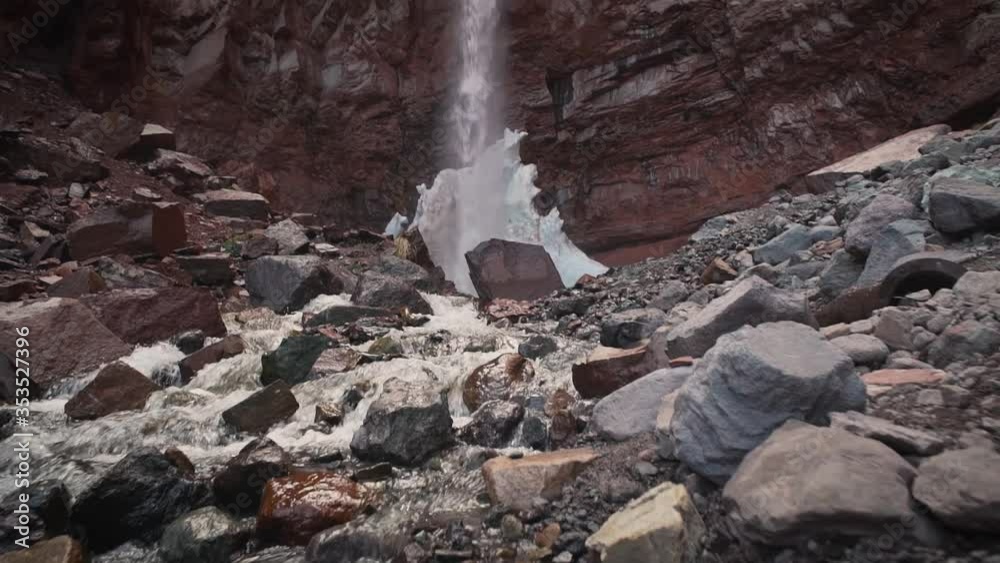 Pan up from rocky ground to large waterfall over red cliffside and blue cloudy sky in slow motion Pan up from rocky ground to large waterfall over red cliffside and blue cloudy sky in slow motion
