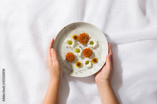 Top view of voile fabric background with female hands holding a plate with floating flowers. 