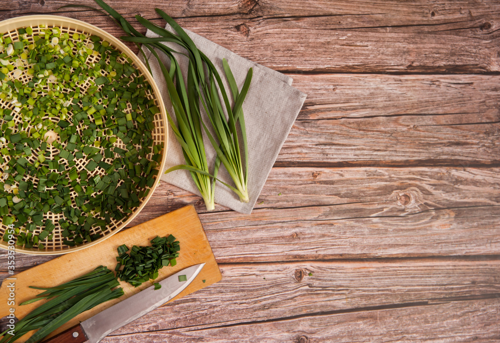 Obraz premium Fresh spicy green garlic feathers are cut with a knife on a cutting Board and placed on a dehydrator grid on a wooden background. Preparation for drying with preservation of vitamins. Free space.