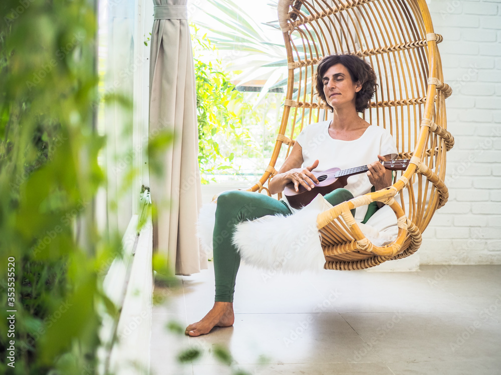 happy and relaxed woman playing ukulele sitting in a bamboo rattan hanging swing chair with green plants