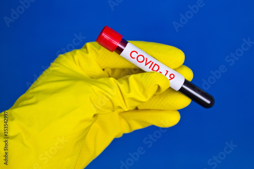 Hand in yellow medical rubber gloves holding test tube with red blood on blue background with the inscription Covid-19.