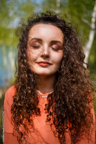 Wood masonry. A girl with long wavy curly hair in an orange guipure dress and shoes on nature, in a forest by the lake, stood near trees and bushes. Young woman smiles and enjoys life