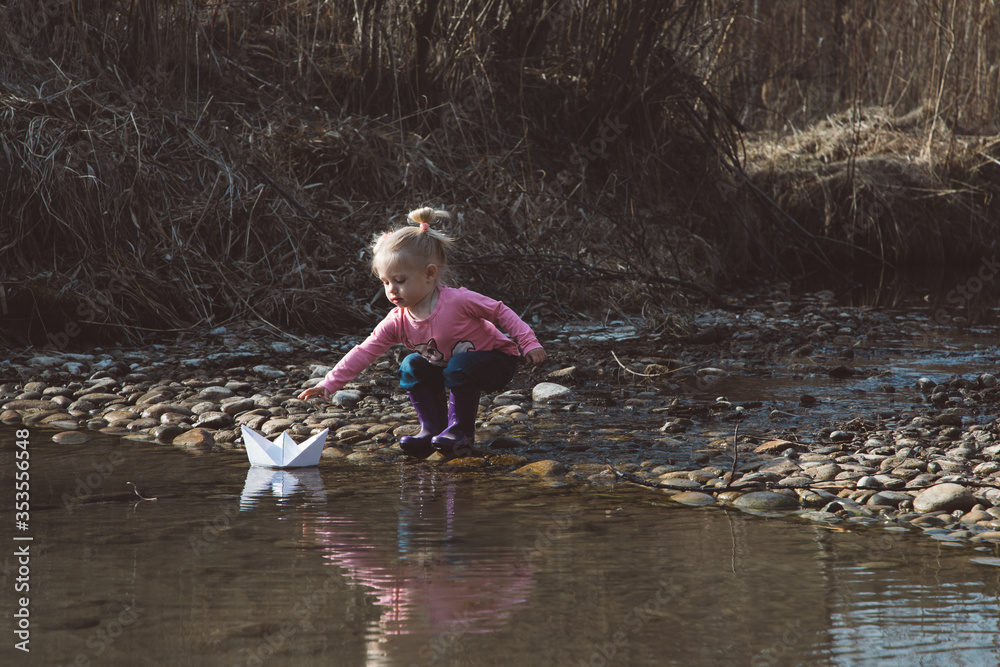 Little girl in rubber boots launches paper white boats in creek in spring or autumn Little girl in rubber boots launches paper white boats in creek in spring or autumn