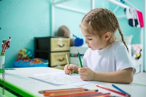 Portrait of a cute little girl looking at the camera and smiling while drawing pictures or doing homework, sitting at a table in the home interior, copy space