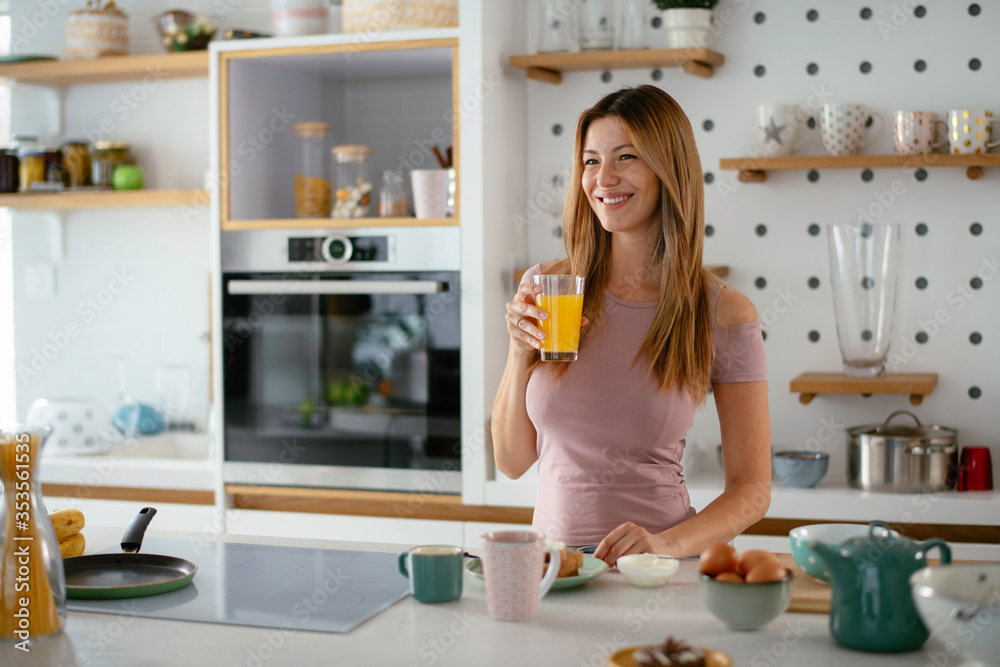  Beautiful woman drinking orange juice in the kitchen. Young woman reading the news online.
