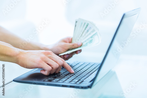 Close-up of person's hands paying online using a laptop while holding US dollars money. Online shopping, banking and e-commerce concept. 