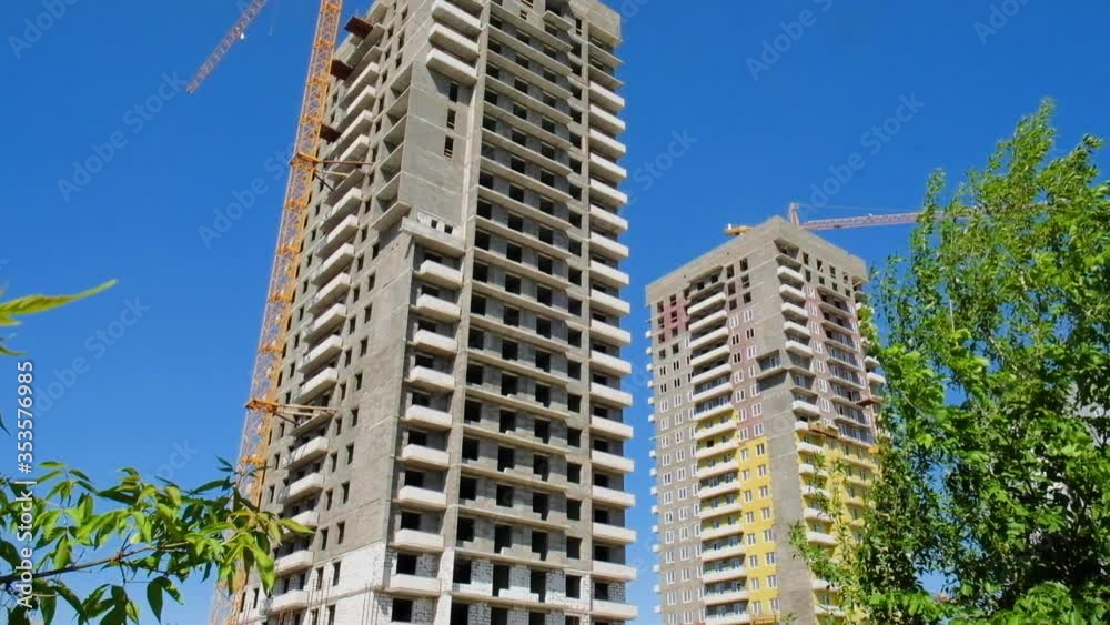 The construction of several multi-story residential buildings of concrete in the city on a summer day against a blue sky and white clouds next to a shady alley. The construction of several multi-story residential buildings of concrete in the city on a summer day against a blue sky and white clouds next to a shady alley.