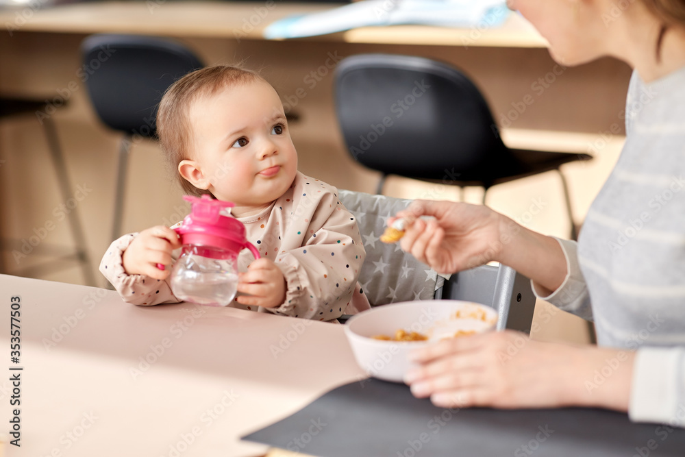 family, eating and people concept - happy mother with puree and spoon feeding little baby girl sitting in highchair at home family, eating and people concept - happy mother with puree and spoon feeding little baby girl sitting in highchair at home