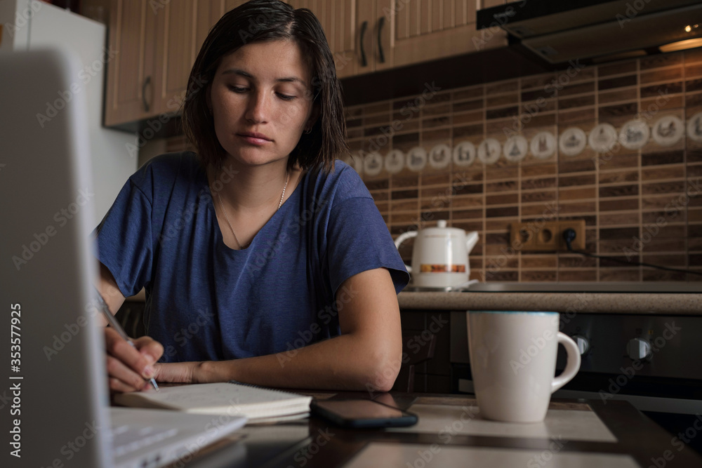 Serious beautiful young adult asian woman taking notes while sitting in front of laptop computer on kitchen table near window
