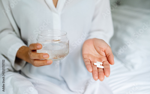 Closeup image of a sick woman holding white pills and a glass of water while sitting on a bed
