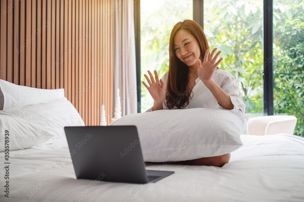 Closeup image of a beautiful asian woman waving hands while using laptop for video call while sitting on a bed at home Closeup image of a beautiful asian woman waving hands while using laptop for video call while sitting on a bed at home