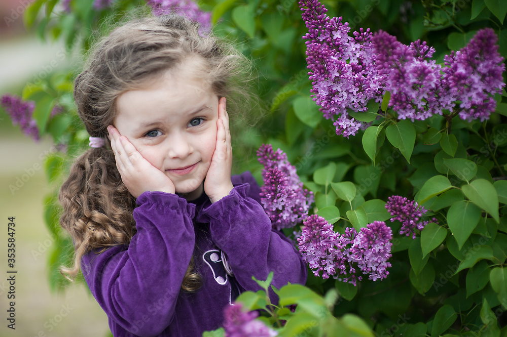 Cute toddler girl with loose curly hair among the bushes of lilac with bunches of flowers Cute toddler girl with loose curly hair among the bushes of lilac with bunches of flowers