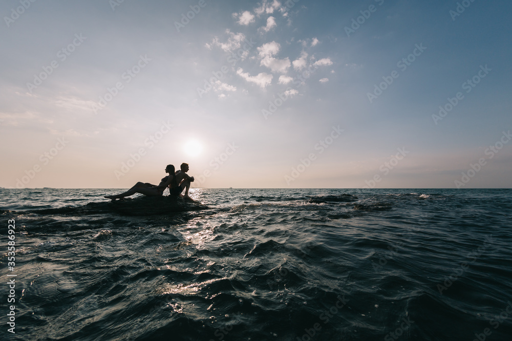 Young couple guy with a girl sitting on a rock washed by the sea on a sunset background