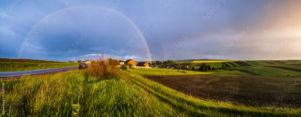 Obraz premium Spring rapeseed and small farmlands fields after rain evening view, cloudy pre sunset sky with colorful rainbow and rural hills. Natural seasonal, climate, farming, countryside beauty concept scene.