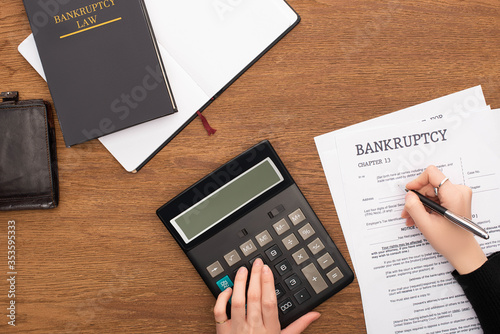 Partial view of woman filling in bankruptcy form and using calculator on wooden background photo