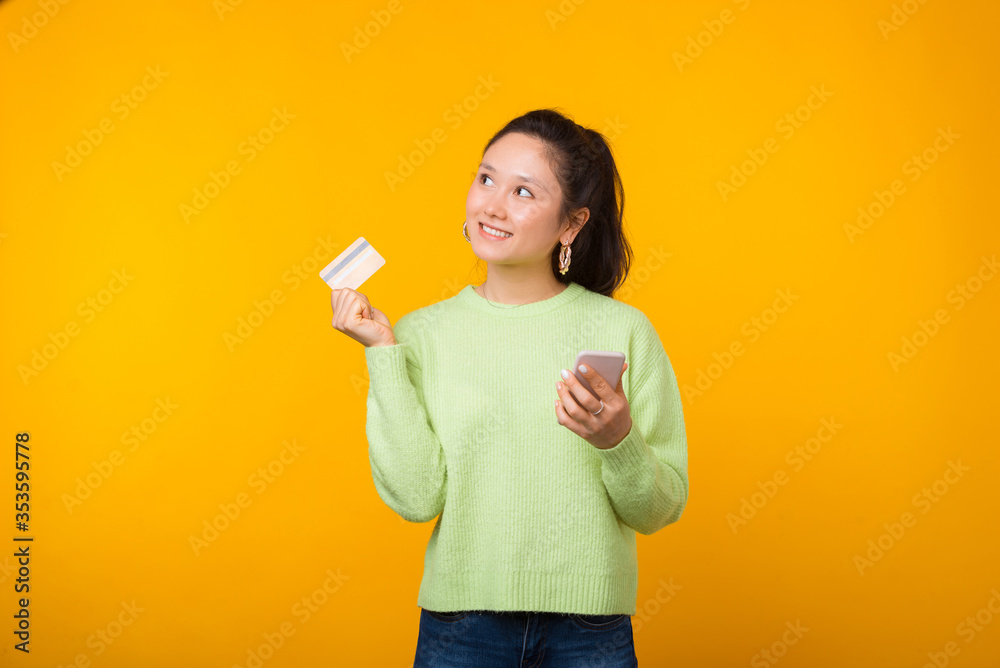 Playful young woman is holding a credit or debit card and her phone while standing on yellow background Playful young woman is holding a credit or debit card and her phone while standing on yellow background