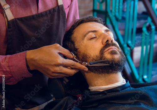 Beard styling. Barber shaping beard of handsome man using a trimmer. Skilled barber trimming beard to his client