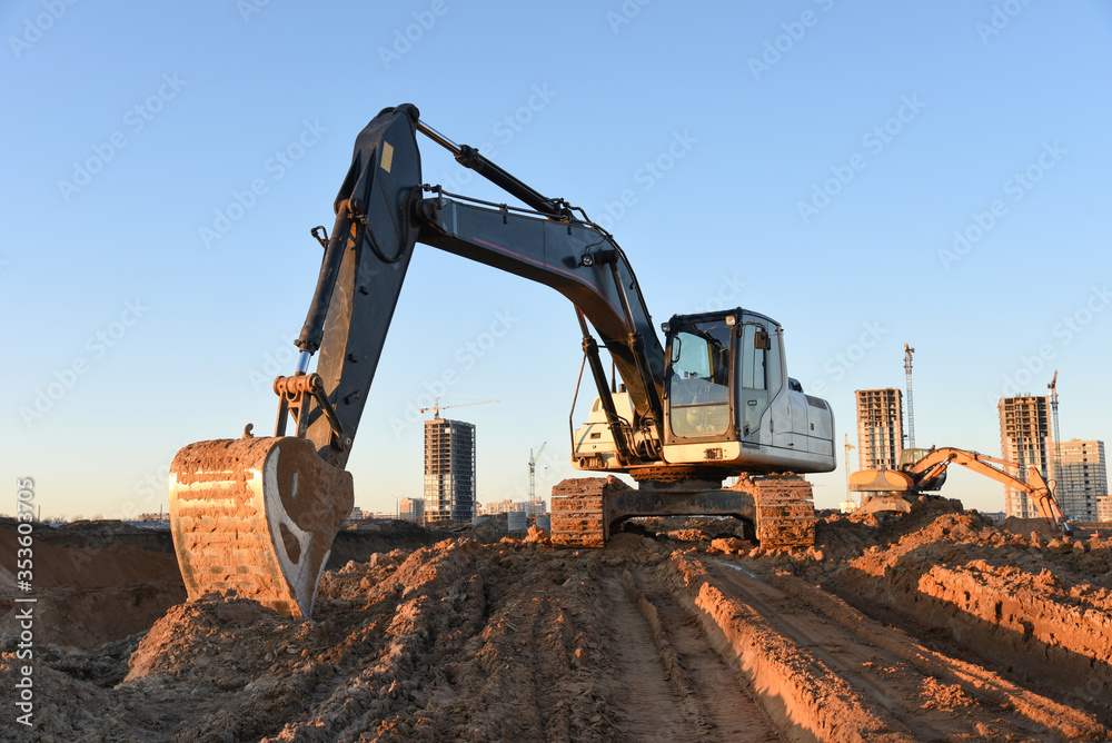 Excavator working at construction site on earthworks. Backhoe digging building foundation. Paving out sewer line. Heavy machinery for road work, excavating, loading, lifting and hauling of cargo Excavator working at construction site on earthworks. Backhoe digging building foundation. Paving out sewer line. Heavy machinery for road work, excavating, loading, lifting and hauling of cargo