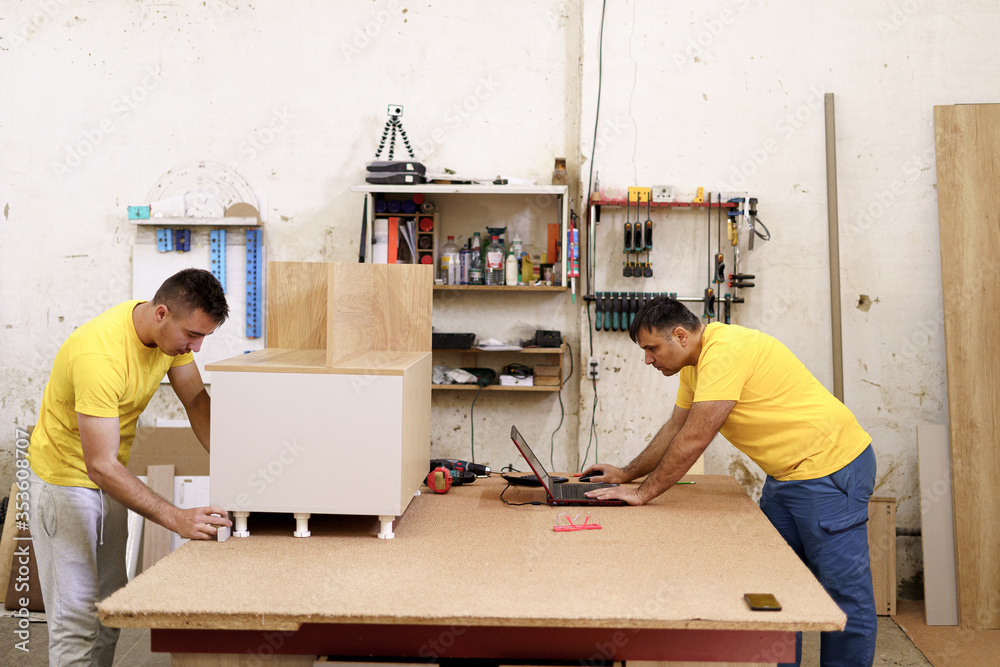 Two carpenters in yellow T-shirts using laptop while discussing furniture in workshop while working with wood together with instruments on the wall in the background Two carpenters in yellow T-shirts using laptop while discussing furniture in workshop while working with wood together with instruments on the wall in the background