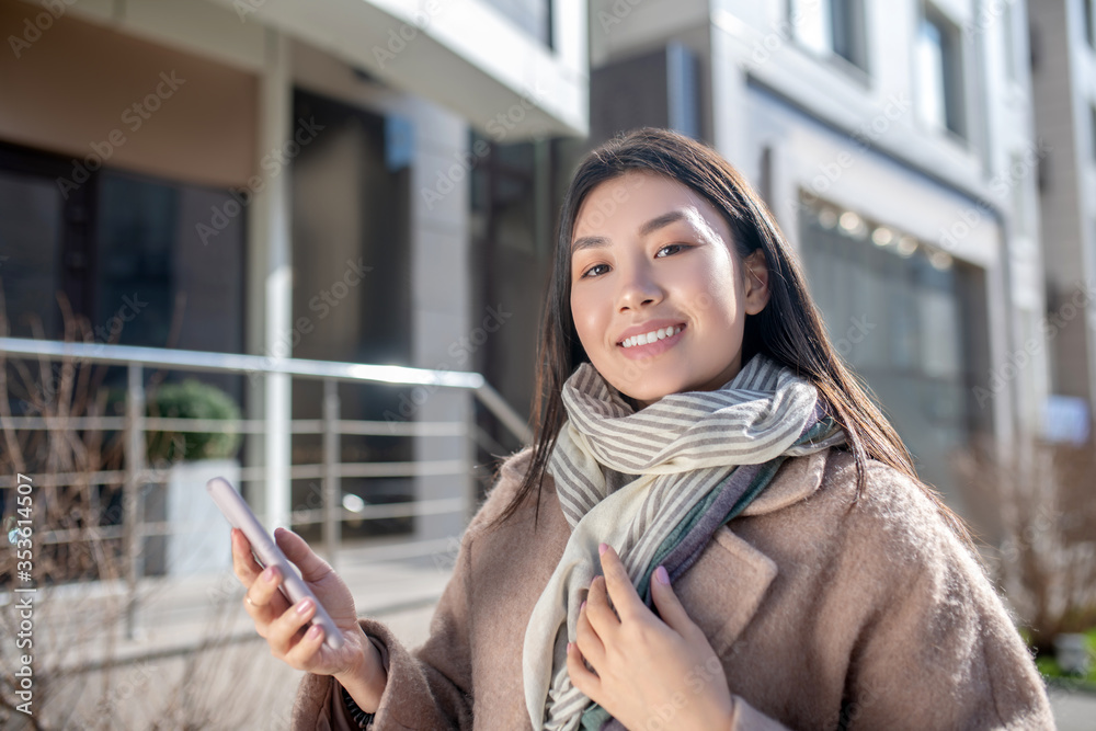 Brunette female in beige coat standing outside, checking her phone, smiling