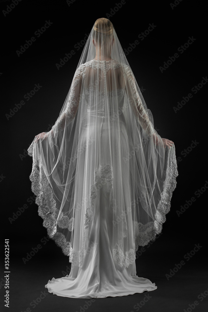 Bride in a beaituful flowing white wedding dress and long lace bridal veil, back view. Black background