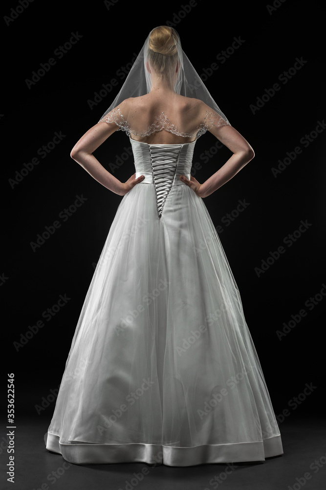 Fototapeta premium Bride in a beaituful long white wedding dress and lace bridal veil, back view. Black background