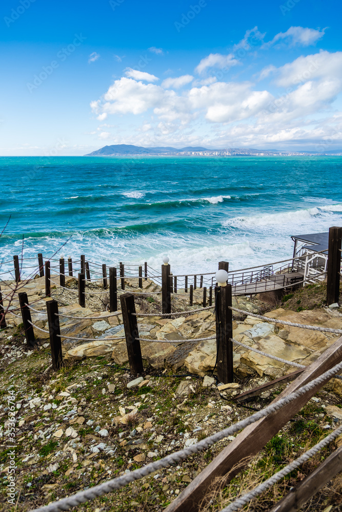 A staircase with stone steps and rope handrails leads down to the green stormy sea. On the horizon mountains and ships on the roadstead of Novorossiysk