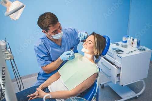 Dentist examining a patient s teeth using dental equipment in dentistry office. Stomatology and health care concept. Young handsome male doctor in disposable medical facial mask  smiling happy woman.