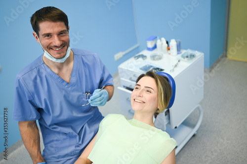 Dentist examining a patient s teeth using dental equipment in dentistry office. Stomatology and health care concept. Young handsome male doctor in disposable medical facial mask  smiling happy woman.