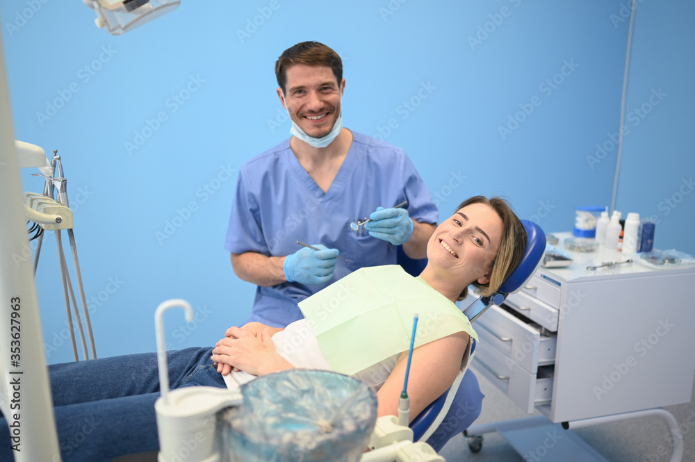 Fototapeta premium Dentist examining a patient's teeth using dental equipment in dentistry office. Stomatology and health care concept. Young handsome male doctor in disposable medical facial mask, smiling happy woman.