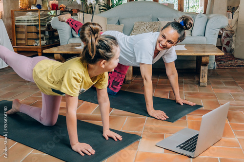 Mother and daughter practicing online yoga classes at home during the quarantine during the coronavirus pandemic. Family doing sports together online from home during isolation. Beautiful young woman 