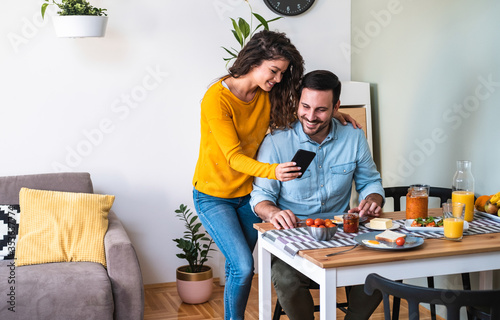 Happy beautiful young couple having breakfast at the kitchen table, looking at mobile phone stock photo