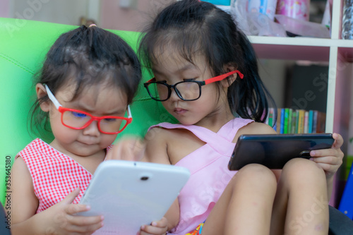 Two kindergarten Asian girls are playing phones and tablets with fun.