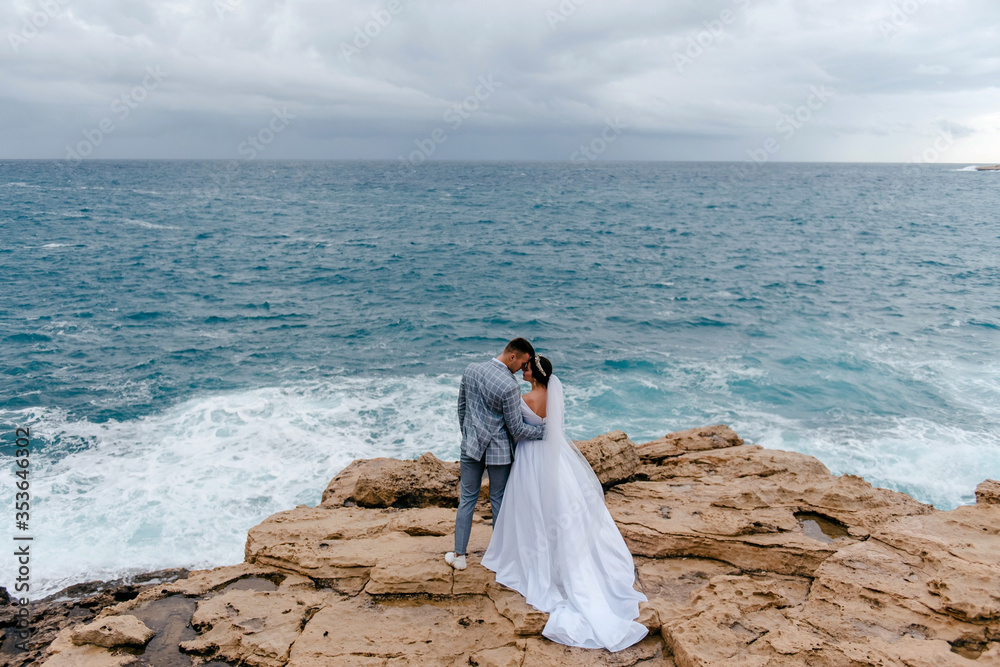 The newlyweds gently hug on the rocks by the sea and enjoy the nature of Cyprus. Rear view. Honeymoon of the newlyweds The newlyweds gently hug on the rocks by the sea and enjoy the nature of Cyprus. Rear view. Honeymoon of the newlyweds