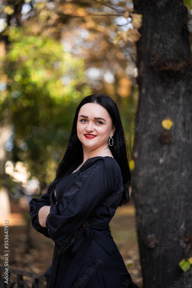 Autumn fashion outdoor. The girl with red lips in fashionable stylish black dress, autumnal lifestyle on the background of blurry yellow-green trees in the park. Vertical Autumn fashion outdoor. The girl with red lips in fashionable stylish black dress, autumnal lifestyle on the background of blurry yellow-green trees in the park. Vertical