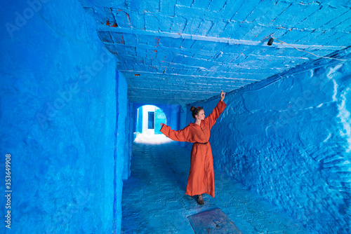 in the long corridor of the blue city of Morocco stands a girl in a long orange dress with her arms raised up, as if dancing © nelen.ru