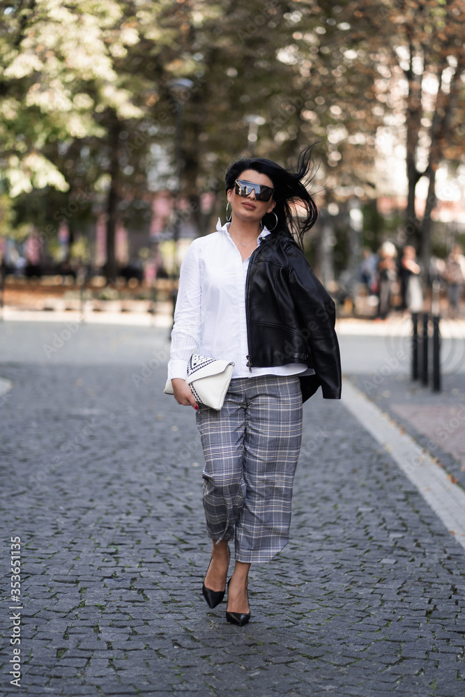 Naklejka premium Autumn fashion outdoor. The girl in fashionable stylish white shirt , pants, jacket and sunglasses, autumnal lifestyle on the background of blurry yellow-green trees in the park. Vertical