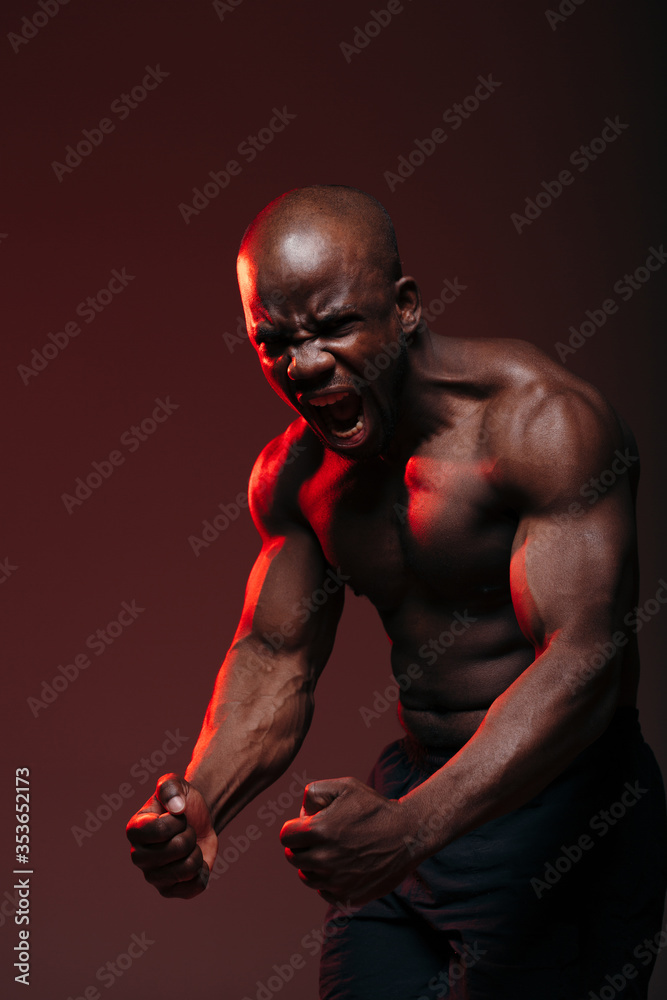 Vertical photo with red light. Muscular african american fighter showing naked torso muscles and screaming.