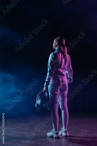 Back view of fencer holding fencing mask and rapier on black background with smoke