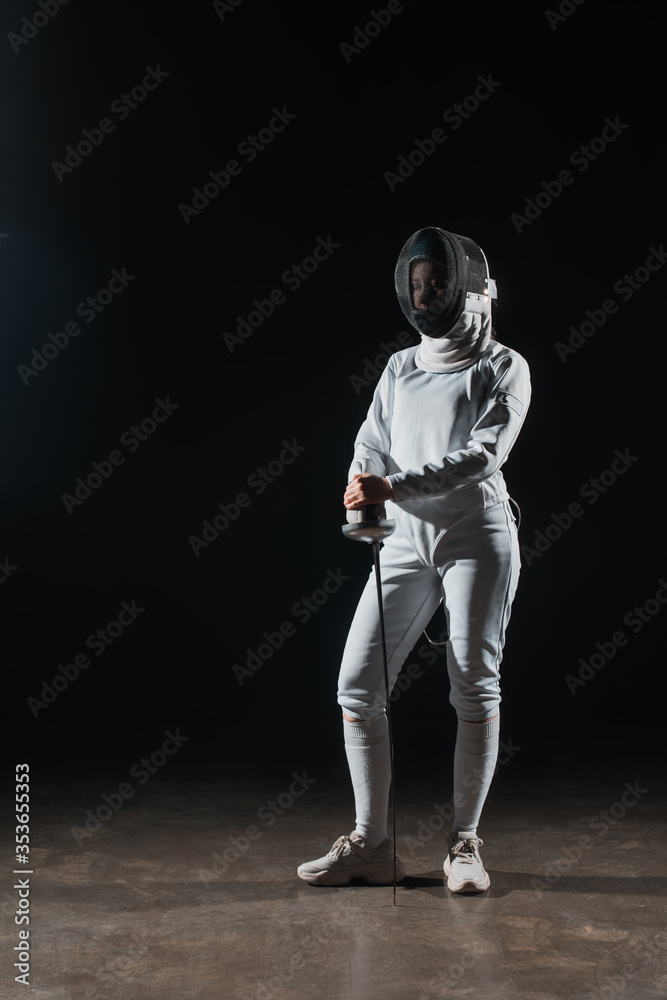 Fencer in fencing mask and suit holding rapier on black background