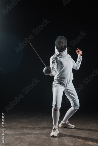 Fencer in fencing mask holding rapier while training under spotlight on black background