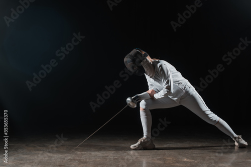 Fencer in fencing mask and suit holding rapier and doing lunge on black background © LIGHTFIELD STUDIOS