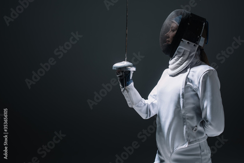 Fotografía Side view of fencer in fencing mask holding rapier isolated on black