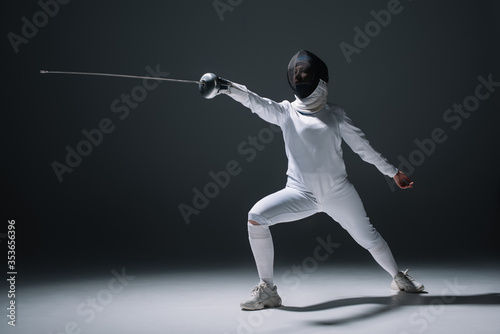 Swordswoman in fencing mask and suit holding rapier on white surface on black background